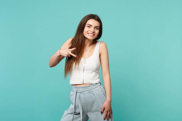 Retrato de mulher jovem sorridente com roupas leves casuais, olhando a câmera, mostrando o sinal da vitória isolado no fundo da parede azul turquesa. emoções sinceras de pessoas, conceito de estilo de vida. simule o espaço da cópia.