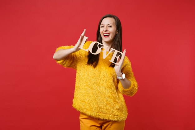 Retrato de mulher jovem sorridente com suéter de pele amarela segurando palavras de madeira letras amor isolado no fundo da parede vermelha brilhante no estúdio. emoções sinceras de pessoas, conceito de estilo de vida. simule o espaço da cópia.