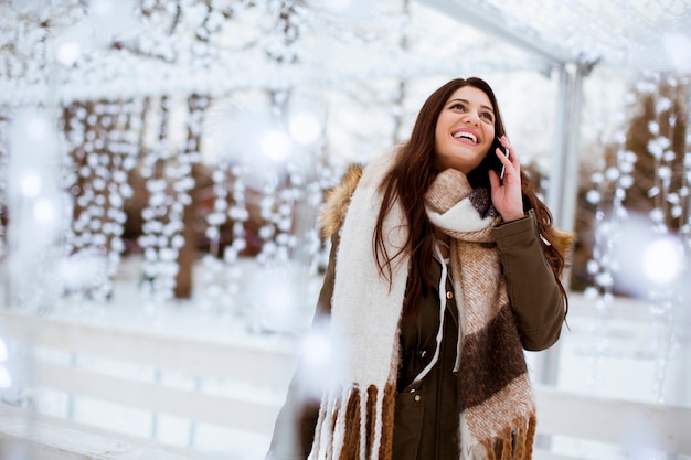 Retrato de mulher jovem sorridente com telefone celular no inverno ao ar livre no mercado de natal