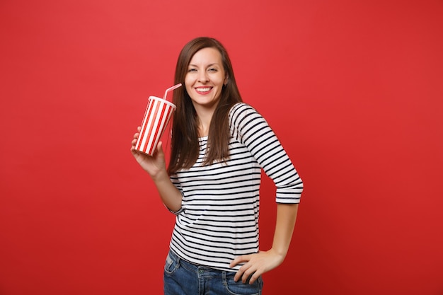 Retrato de mulher jovem sorridente em roupas listradas casuais, segurando um copo plástico de coca-cola ou refrigerante isolado no fundo da parede vermelha brilhante. Emoções sinceras de pessoas, conceito de estilo de vida. Simule o espaço da cópia.