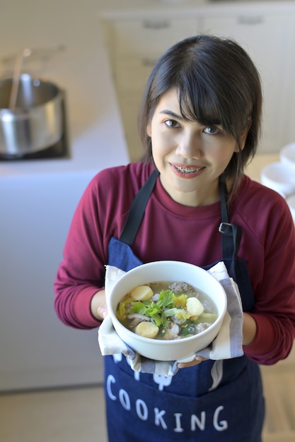 Retrato de mulher jovem sorridente na cozinha moderna e segurando uma tigela de sopa com tofu