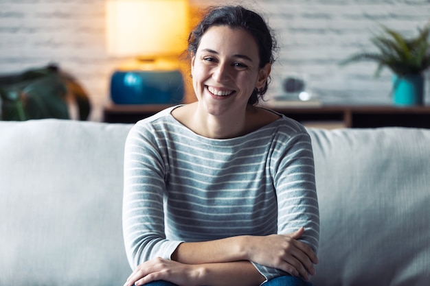 Retrato de mulher jovem sorridente, olhando para a câmera enquanto está sentado no sofá da sala de estar em casa.