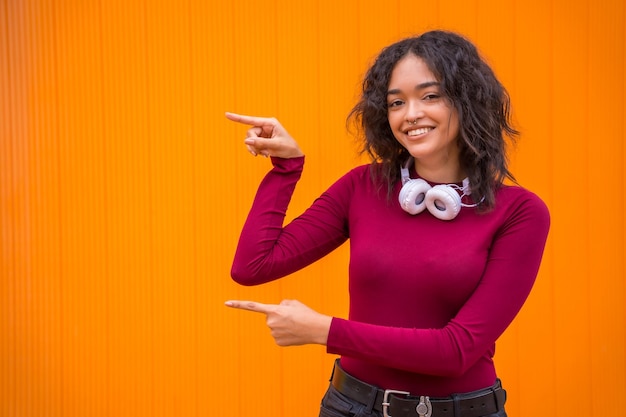 Retrato de mulher latina com fones de ouvido sorrindo conceito de tecnologia em um fundo laranja apontando para o espaço esquerdo da cópia
