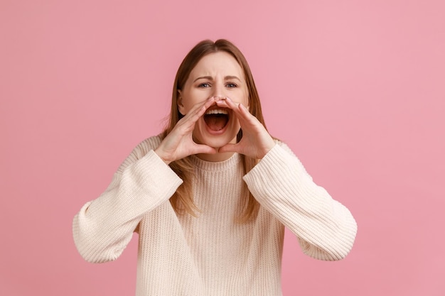 Retrato de mulher loira adulta jovem agressiva em pé segurando os braços perto da boca aberta e gritando alto vestindo suéter branco Tiro de estúdio interior isolado no fundo rosa