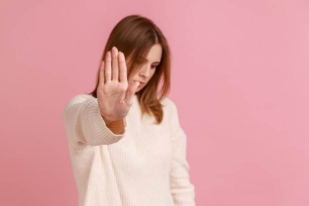 Retrato de mulher loira adulta jovem infeliz em pé com gesto de parada olhando para longe cuidado para não tocar vestindo suéter branco Tiro de estúdio interno isolado no fundo rosa