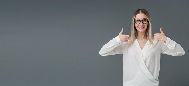 Retrato de mulher loira bonita usando óculos elegantes com sorriso caloroso e ela está confiante Ela gesticula polegares para cima Inclui espaço de cópia