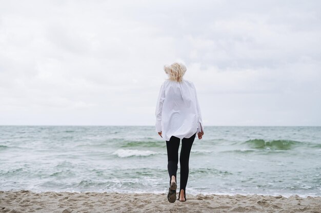 Foto retrato de mulher loira elegante de camisa branca em uma praia de areia em um mar de tempestade em um tempo ventoso