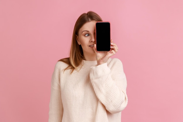 Retrato de mulher loira engraçada cobrindo o olho com o celular com tela vazia para propaganda, olhando para longe, vestindo suéter branco. tiro de estúdio interior isolado no fundo rosa.