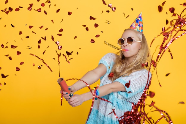 Foto retrato de mulher loira engraçada com chapéu de aniversário e confete vermelho na celebração de fundo amarelo e ...