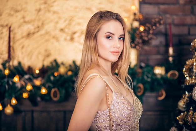 Retrato de mulher loira luxuosa em vestido de noite dourado na árvore de Natal e fundo de velas. Sexy linda garota sorri e olha para baixo. Feriados, celebração e conceito de pessoas