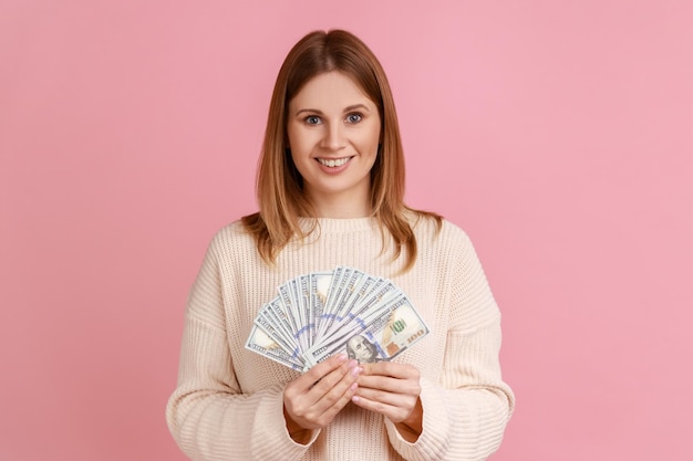Retrato de mulher loira rica feliz segurando grande fã de notas de dólar olhando para a câmera com expressão positiva vestindo suéter branco Tiro de estúdio interior isolado no fundo rosa