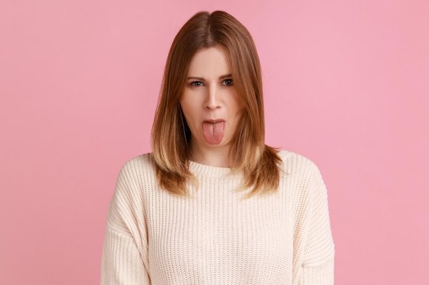 Retrato de mulher loira saindo da língua e olhando para a câmera, provocando com expressão impertinente, comportamento desobediente, vestindo suéter branco. Tiro de estúdio interior isolado no fundo rosa.