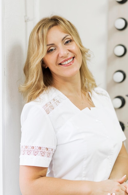 Foto retrato de mulher madura médico cosmetologista dermatologista em um jaleco fica em seu escritório, olha para a câmera e sorri.