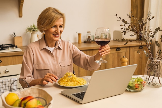 Retrato de mulher madura sênior sorridente segurando o copo de vinho enquanto usa o laptop na mesa da cozinha. Freelance, trabalhando no conceito de casa.