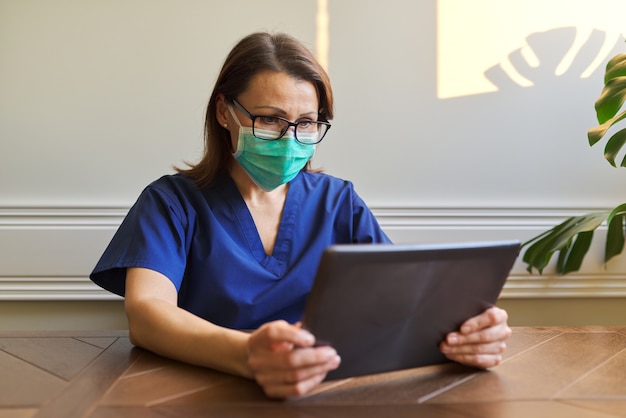 Retrato de mulher médica com máscara protetora médica com tablet digital sentada à mesa no escritório