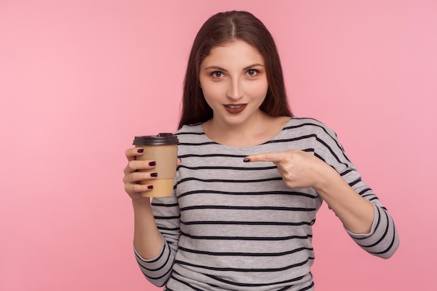Retrato de mulher morena atraente em moletom listrado, apontando para o café e sorrindo para a câmera, bebida com cafeína, aumento de energia na manhã. tiro de estúdio interior isolado no fundo rosa