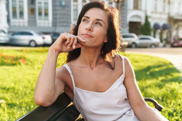 Retrato de mulher morena com vestido de verão pensando e olhando para o lado enquanto está sentado no banco ao ar livre