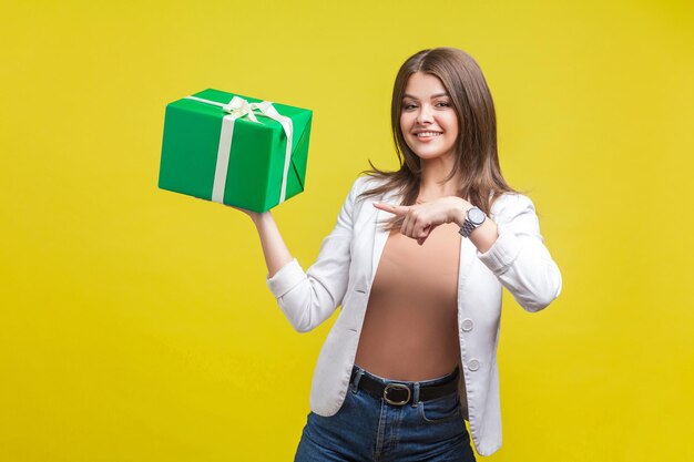 Retrato de mulher morena feliz de jaqueta e jeans, olhando para a câmera com um sorriso fofo e apontando para a caixa de presente verde em sua mão férias presente estúdio interno tiro isolado em fundo amarelo