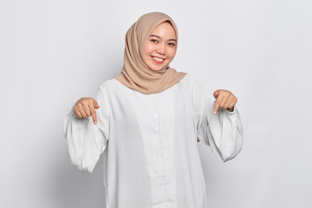 Retrato de mulher muçulmana asiática sorrindo apontando dedos para baixo convidando clientes para um evento especial isolado sobre fundo branco