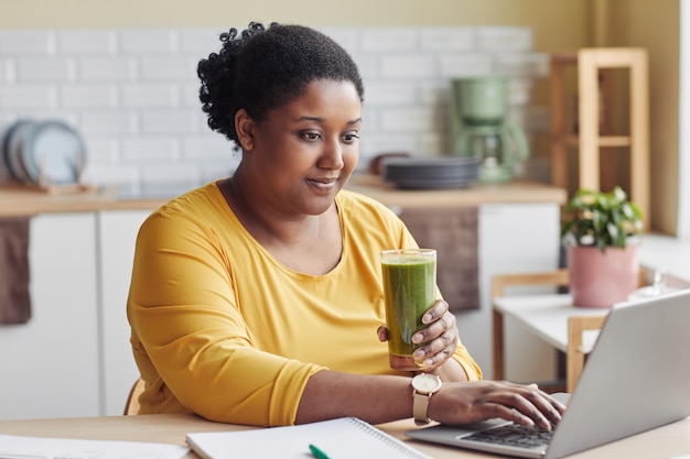 Retrato de mulher negra com excesso de peso bebendo smoothie e usando laptop em casa