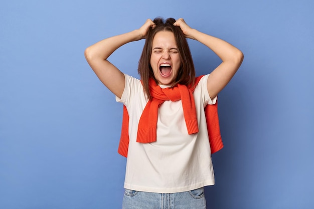 Retrato de mulher nervosa desesperada vestindo roupas de estilo casual em pé isolado sobre fundo azul mantém os olhos fechados puxando o cabelo gritando alto