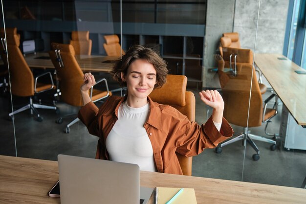 Foto retrato de mulher no local de trabalho, funcionário de escritório nômade digital, sentado na mesa com laptop estica os braços