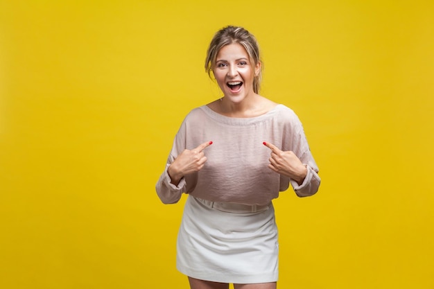 Retrato de mulher orgulhosa espantada com cabelo louro na blusa casual em pé com a boca aberta apontando para si mesma surpresa com a realização de um estúdio interno isolado em fundo amarelo