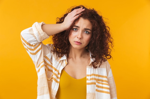 Retrato de mulher perplexa de 20 anos com cabelo encaracolado agarrado à cabeça, isolado em amarelo