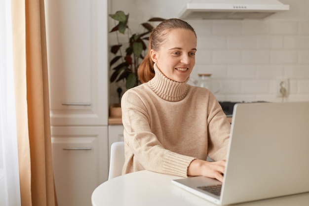 Retrato de mulher positiva sorridente com penteado de rabo de cavalo vestindo suéter bege estilo casual, sentado à mesa na cozinha e trabalhando online, usando pc portátil e internet sem fio.