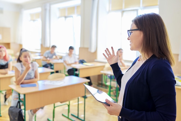 Retrato de mulher professora de meia-idade falando em sala de aula com alunos, filhos adolescentes. Professora com tablet digital ensinando, falando. Educação, escola, faculdade, conceito de ensino