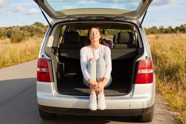 Retrato de mulher relaxada atraente sentada na parte de trás do carro linda fêmea vestindo camisa branca e jeans sentado no porta-malas aberto de um carro com os olhos fechados tomando sol