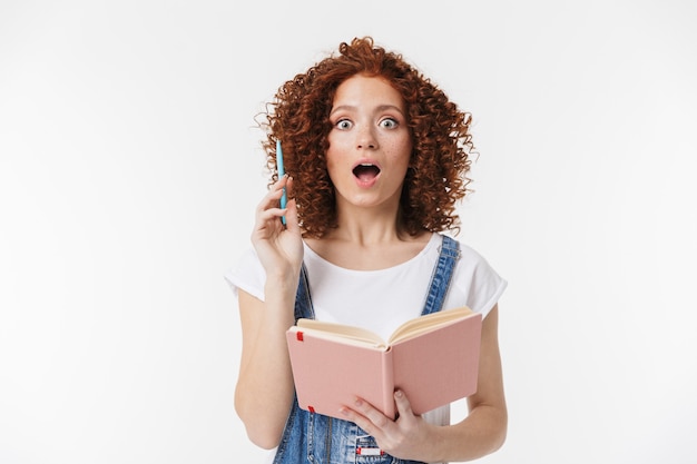 Foto retrato de mulher ruiva encaracolada de 20 anos, surpresa, vestindo macacão jeans, sorrindo e escrevendo notas em seu diário ou planejador do dia isolado sobre a parede branca