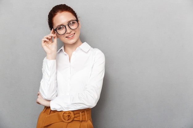 Retrato de mulher ruiva feliz, 20 anos, com roupa formal, segurando óculos e sorrindo no escritório, isolado sobre o cinza