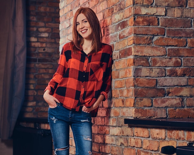 Retrato de mulher ruiva vestida com uma camisa de lã vermelha e jeans sobre a parede de tijolos vermelhos.