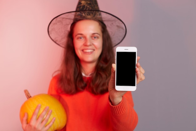 Retrato de mulher segurando grande abóbora laranja madura vestindo suéter e chapéu de bruxa mostrando celular com display vazio com espaço de cópia para anúncio isolado sobre fundo colorido de luz neon