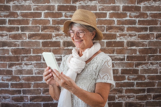 Retrato de mulher sênior atraente em pé de chapéu isolado no fundo da parede de tijolos usando telefone celular em videochamada