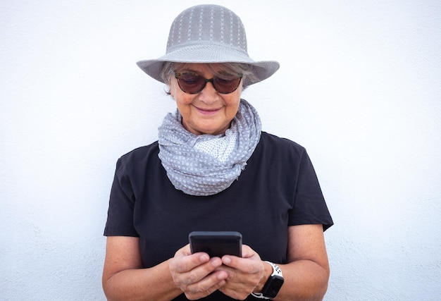 Retrato de mulher sênior atraente em preto com chapéu cinza e cachecol usando telefone celular para mensagens isoladas em fundo branco