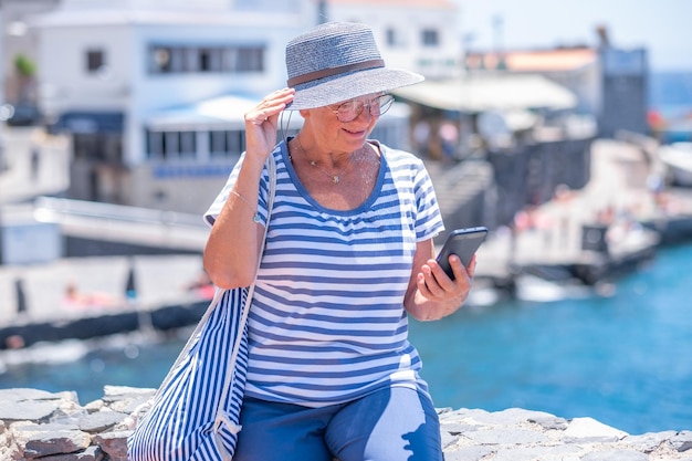 Retrato de mulher sênior caucasiana sorridente atraente lendo uma mensagem no celular enquanto está sentado ao ar livre no mar aproveitando as férias de verão