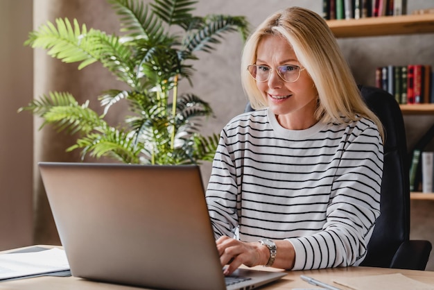 Retrato de mulher sênior em óculos sentado à mesa no escritório e trabalhando no laptop