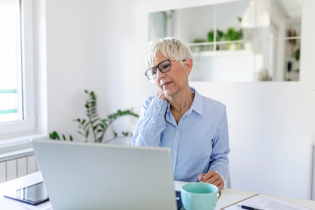 Retrato de mulher sênior estressada, sentada na mesa do escritório em casa em frente ao laptop, tocando o pescoço dolorido com expressão de dor, sofrendo de dor nas costas depois de trabalhar no laptop