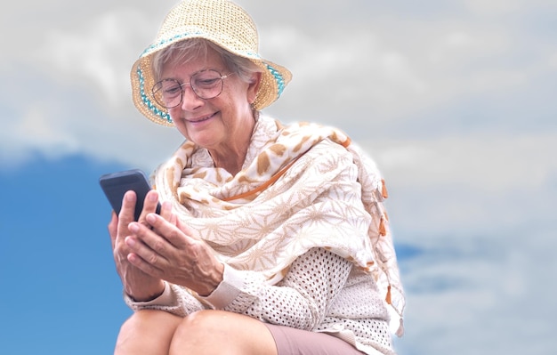 Retrato de mulher sênior sorridente usando óculos e chapéu de verão usando telefone celular sentado ao ar livre desfrutando de liberdade de tecnologia e relaxando Espaço de cópia de céu nublado
