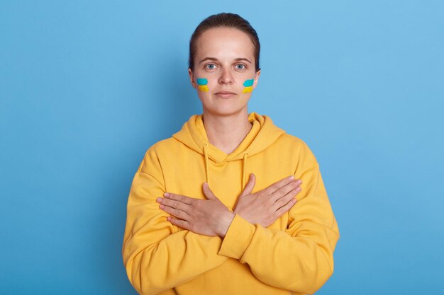 Retrato de mulher séria vestindo capuz com bandeira azul e amarela nas bochechas isoladas sobre fundo azul mantém as mãos cruzadas no peito olhando para a câmera Glória à Ucrânia