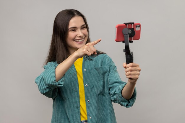 Retrato de mulher sorridente, apontando para a câmera do celular com sorriso fluindo conversando com seguidores usa steadicam vestindo jaqueta estilo casual Tiro de estúdio interno isolado em fundo cinza