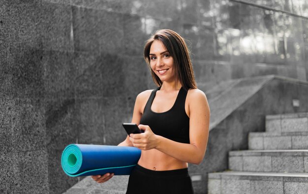 Retrato de mulher sorridente e apta com corpo perfeito, segurando o tapete de ioga dobrado na mão e usando o smartphone