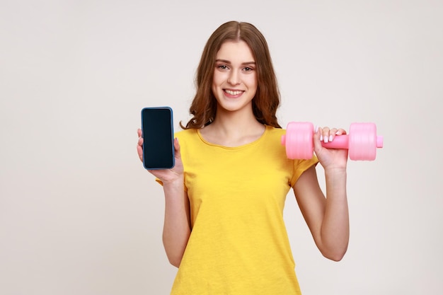 Retrato de mulher sorridente feliz em camiseta casual amarela segurando haltere rosa e smartphone de tela em branco nas mãos satisfeito com o aplicativo de esportes tiro de estúdio interno isolado em fundo cinza