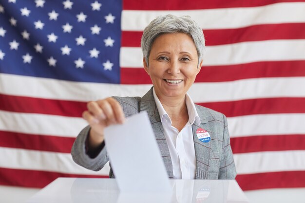 Retrato de mulher sorridente sênior colocando boletim de voto nas urnas e enquanto posava contra a bandeira americana no dia da eleição, copie o espaço