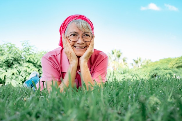 Retrato de mulher sorridente sênior em rosa deitada na grama no parque público tendo momentos relaxados