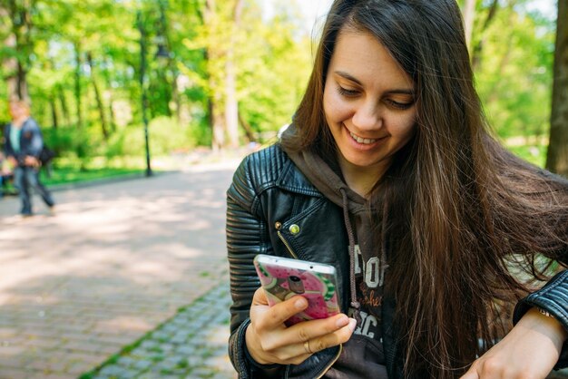 Retrato de mulher sorrindo jovem adulto sentado no banco no parque usando telefone