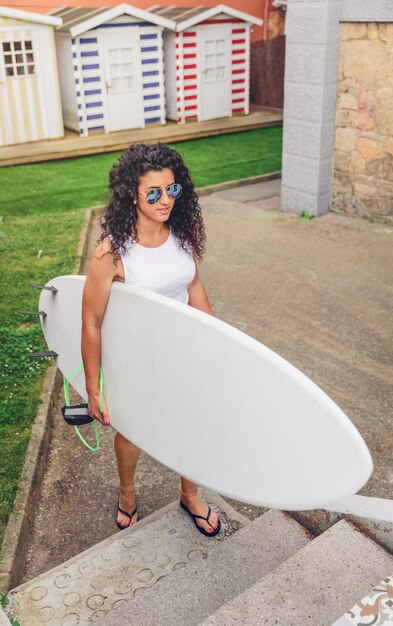 Retrato de mulher surfista morena encaracolada com top branco e óculos escuros andando com uma prancha de surf