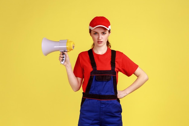 Retrato de mulher trabalhadora mandona estrita segurando o megafone nas mãos, mantém a mão no quadril, olhando para a câmera com expressão séria, vestindo macacão. Tiro de estúdio interior isolado em fundo amarelo.